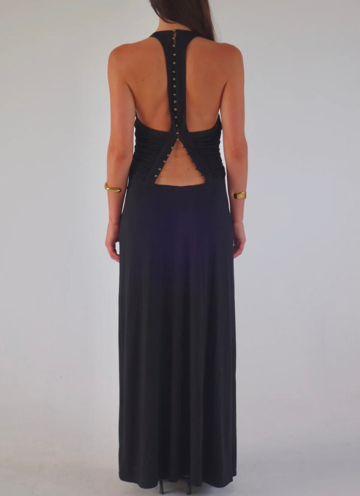 A woman viewed from the back wearing a sleeveless black maxi dress with a deep open back, buttoned along the straps, and a long flowing skirt against a plain light gray studio background.