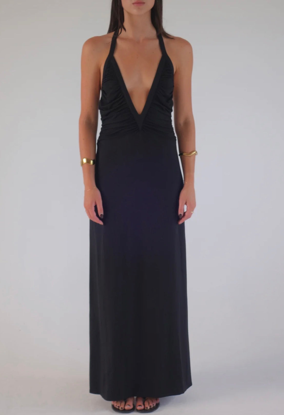 A woman modeling a black halter-neck maxi dress with a deep plunging neckline and gathered ruching at the bust, shown against a plain light gray studio background.