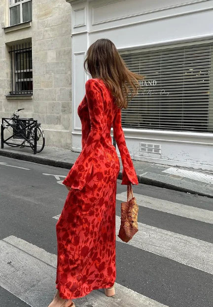 A woman stands on a city crosswalk wearing a long-sleeved red maxi dress with a tonal red-and-black leopard print. She is holding a brown woven handbag and photographed from behind against a street backdrop with a stone building and window blinds.
