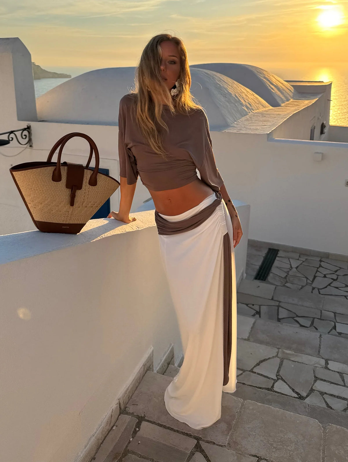 A woman posing on a white-walled terrace at sunset wearing a taupe crop top and a white maxi skirt with a taupe side panel, next to a woven straw handbag.