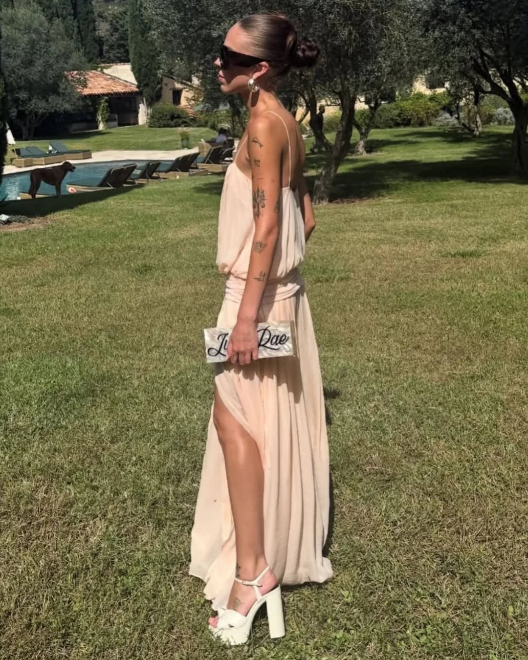 A woman stands on a grassy lawn wearing a draped, sleeveless maxi dress in a light blush pink or beige tone, with a high slit, white platform sandals, and a small white handbag. Trees, a swimming pool, and patio chairs are visible in the background.
