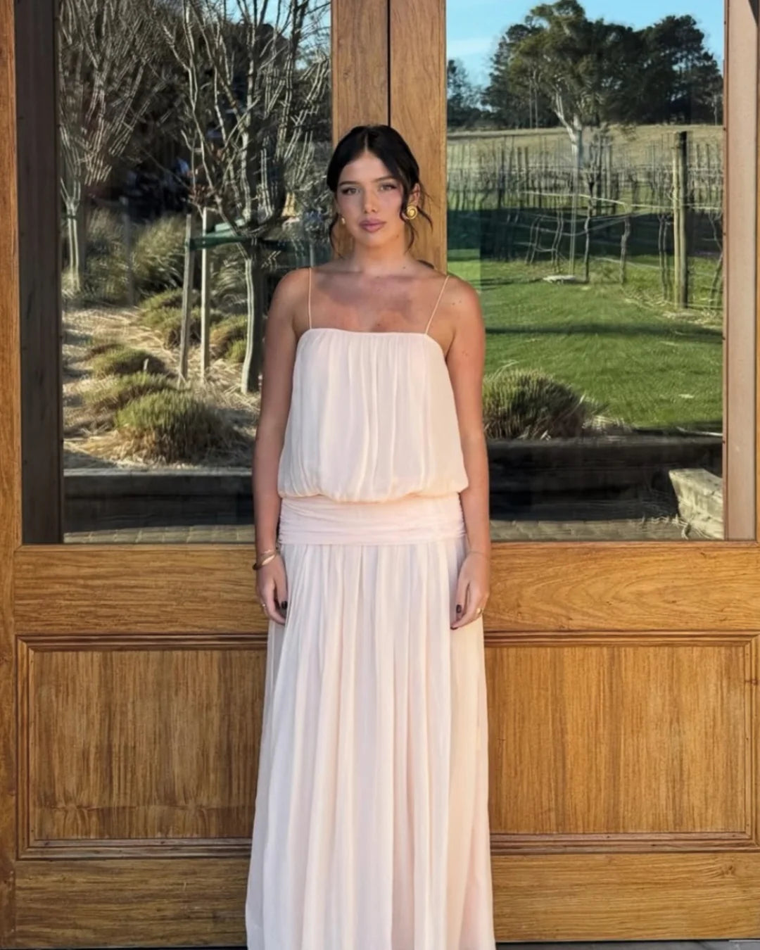 A woman wearing a light peach chiffon strapless maxi dress stands in front of a large wooden door with a vineyard visible in the background.