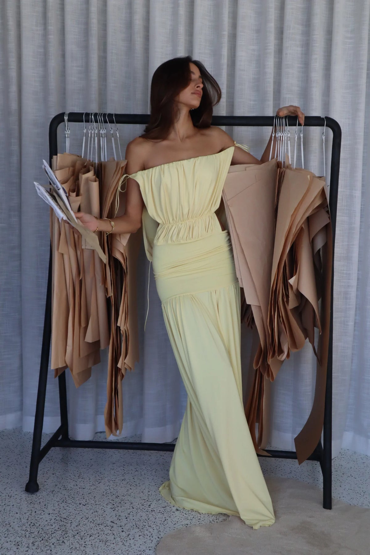 A woman stands in front of a clothing rack wearing an off-the-shoulder pale yellow maxi dress, holding fabric swatches in a studio setting with a light gray curtain backdrop.