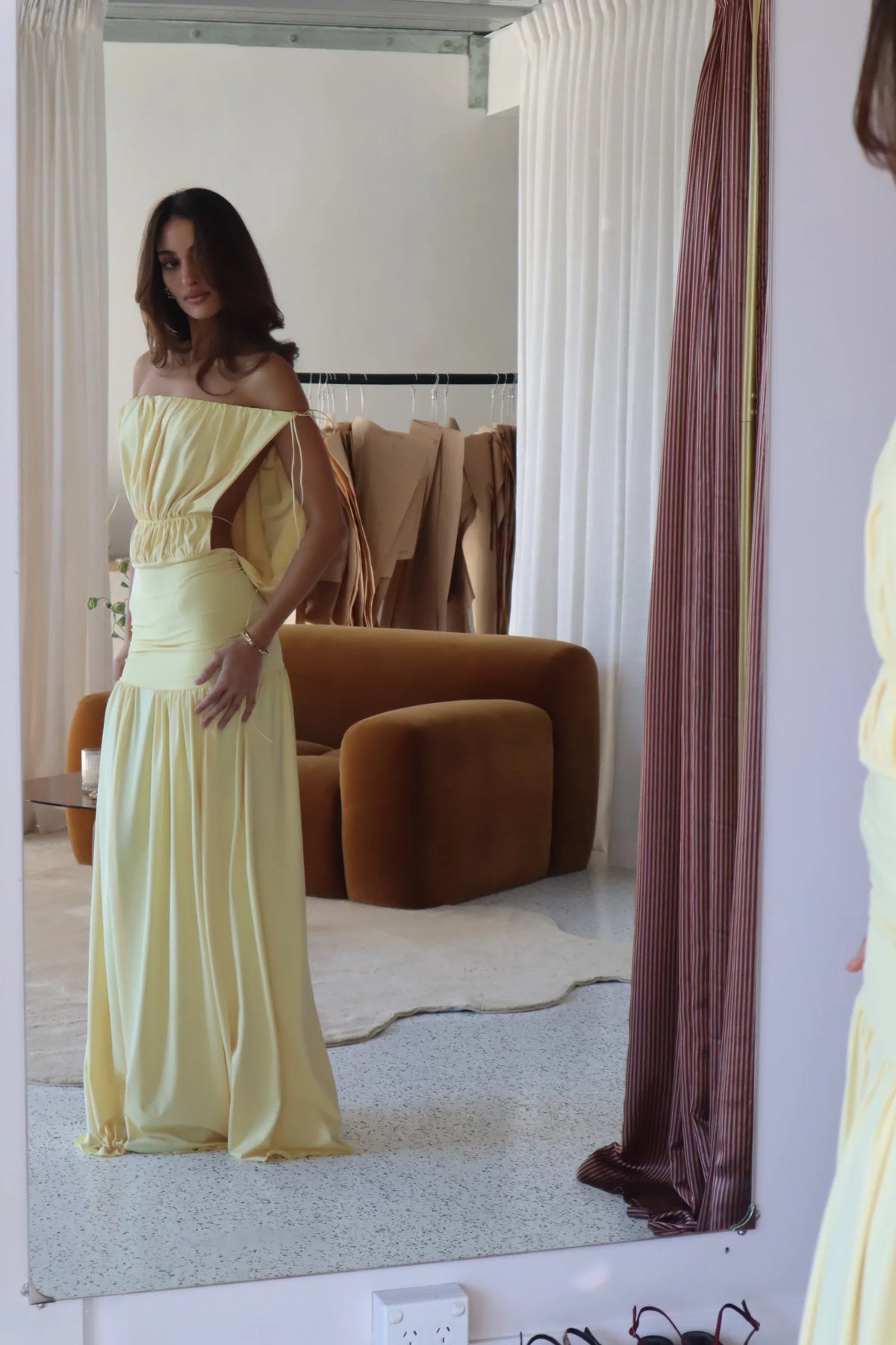  Model wearing a pale yellow off-the-shoulder maxi dress with a gathered bodice, fitted drop waist and flowing skirt, photographed in front of a mirror.

