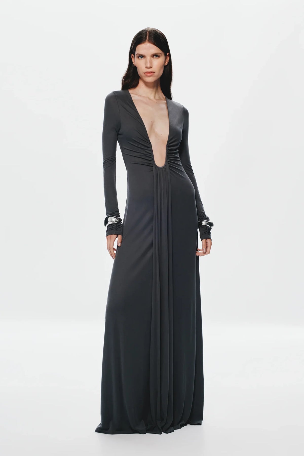 A model wearing a floor-length, dark charcoal gray gown with long sleeves, a deep plunging neckline, and gathered detailing on the bodice, set against a plain light gray studio background.