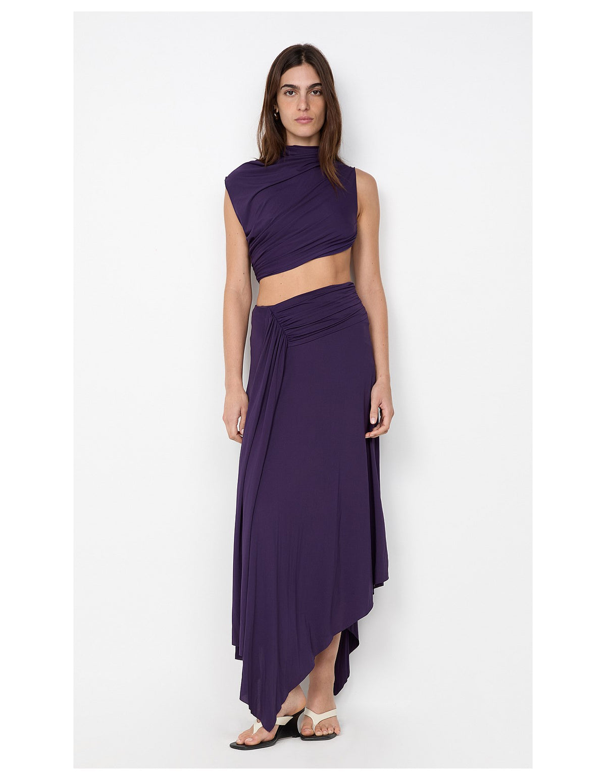  Model wearing a deep plum two-piece set featuring an asymmetrical draped crop top and matching ruched maxi skirt with a flowing asymmetric hem.
