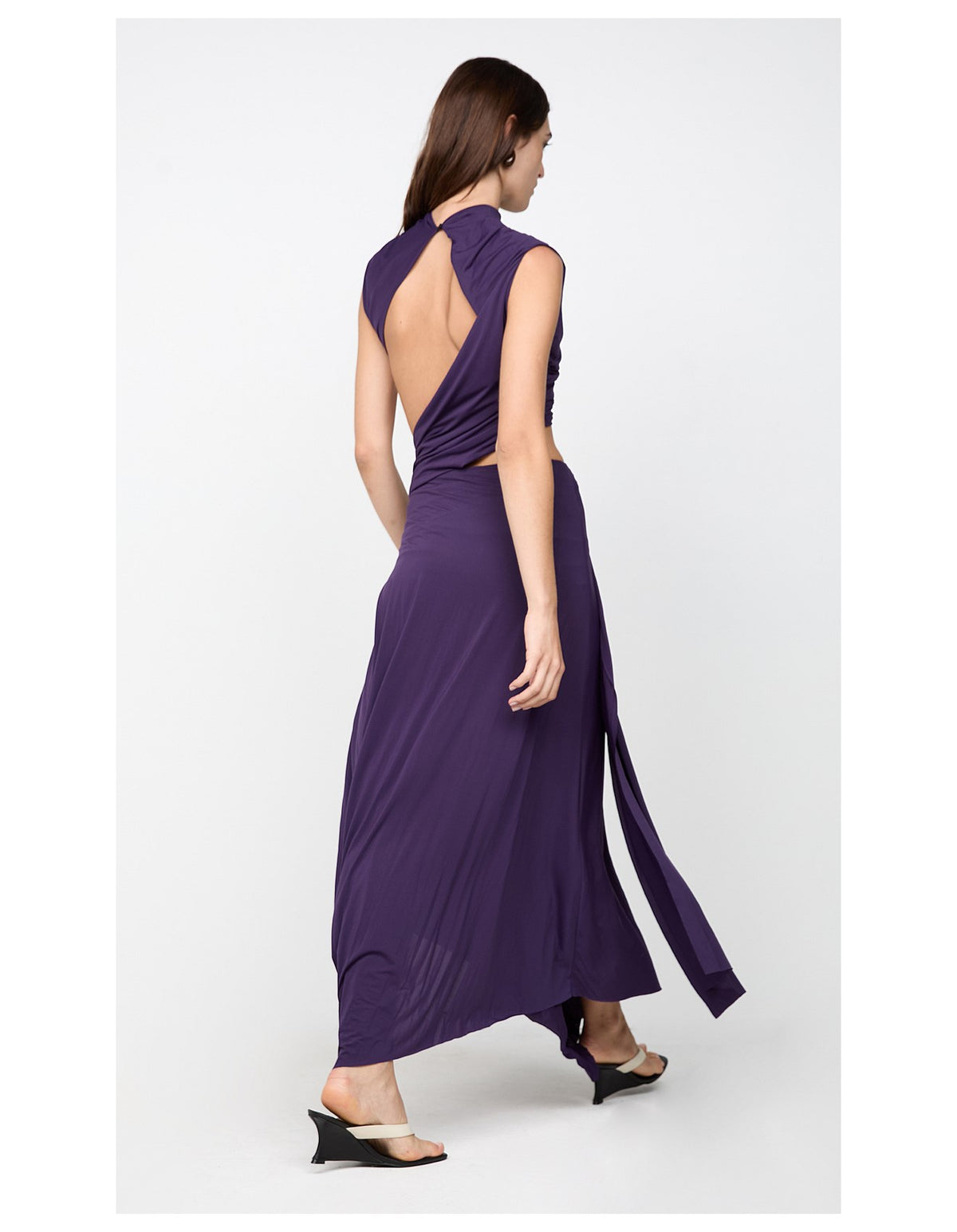 A woman modeling a sleeveless, high-neck, open-back maxi dress in deep purple, shown from the back against a light gray studio background.