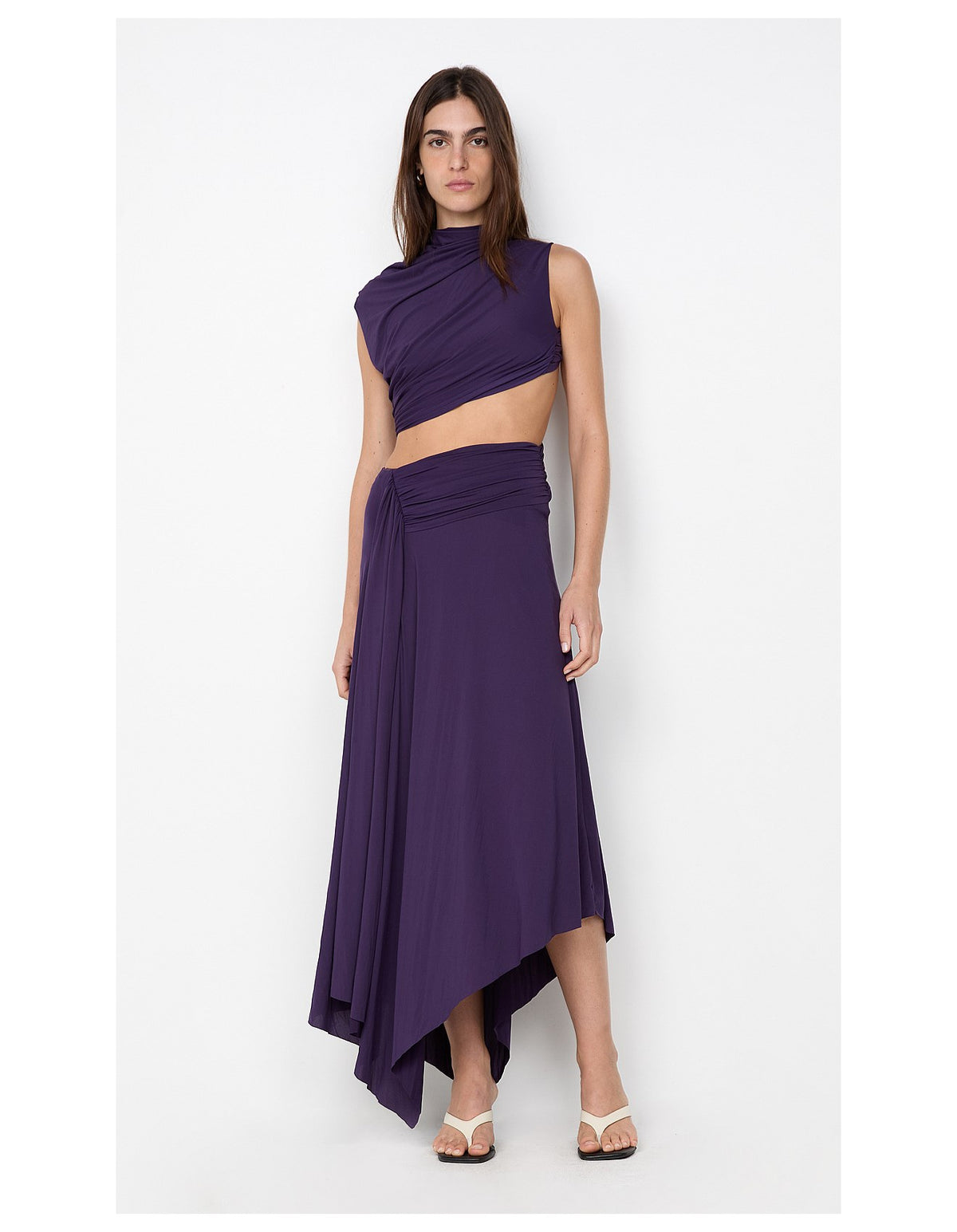  Model wearing a deep plum two-piece set featuring an asymmetrical draped crop top and matching ruched maxi skirt with a flowing asymmetric hem.
