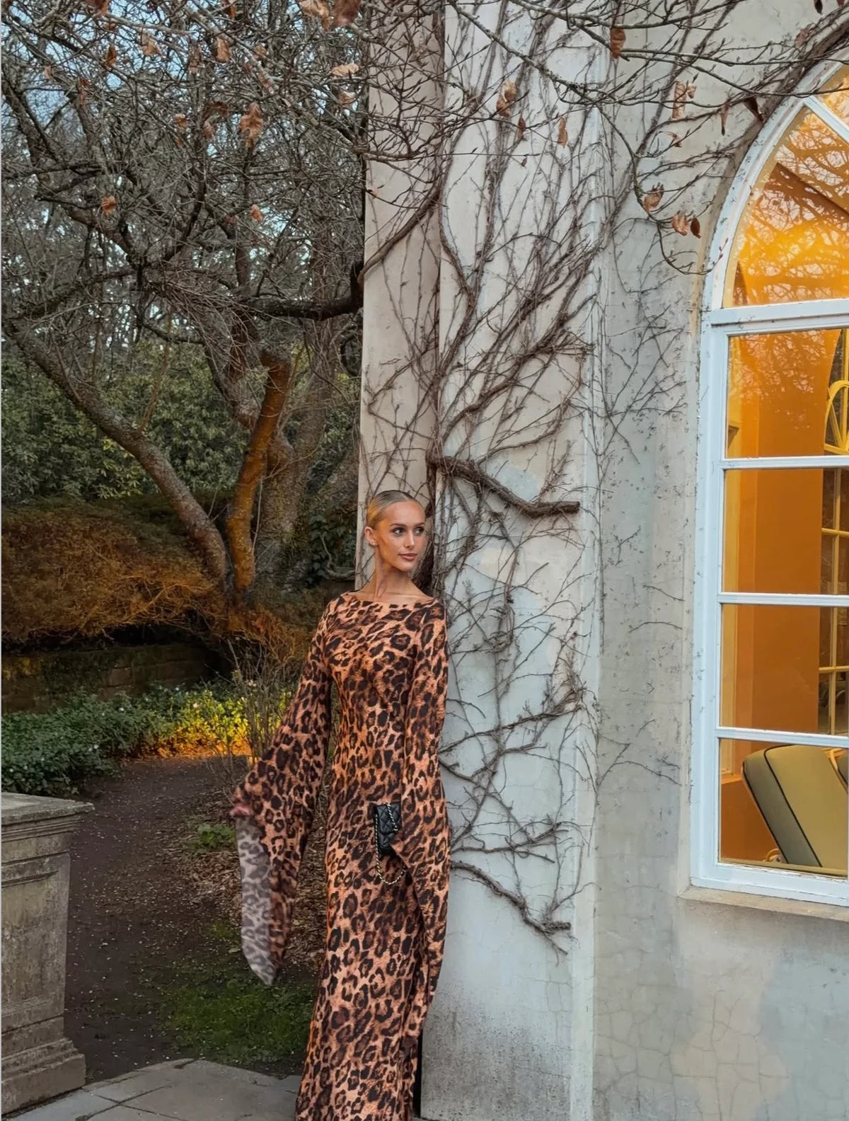 Model wearing the Helsa Roslind leopard print maxi dress with a fitted floor-length silhouette and dramatic draped sleeves, photographed outdoors beside an ivy-covered building.
