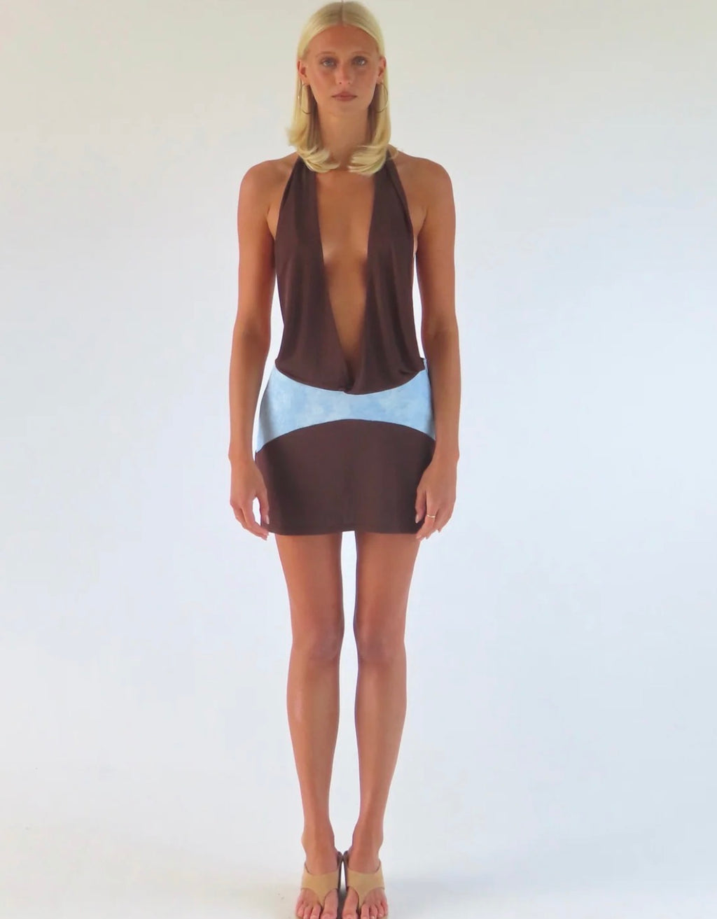  Model wearing the Sola Musa Oma Mini Dress in Bruna, featuring a deep V brown halter neckline, open silhouette and contrasting sky blue curved drop-waist panel.
