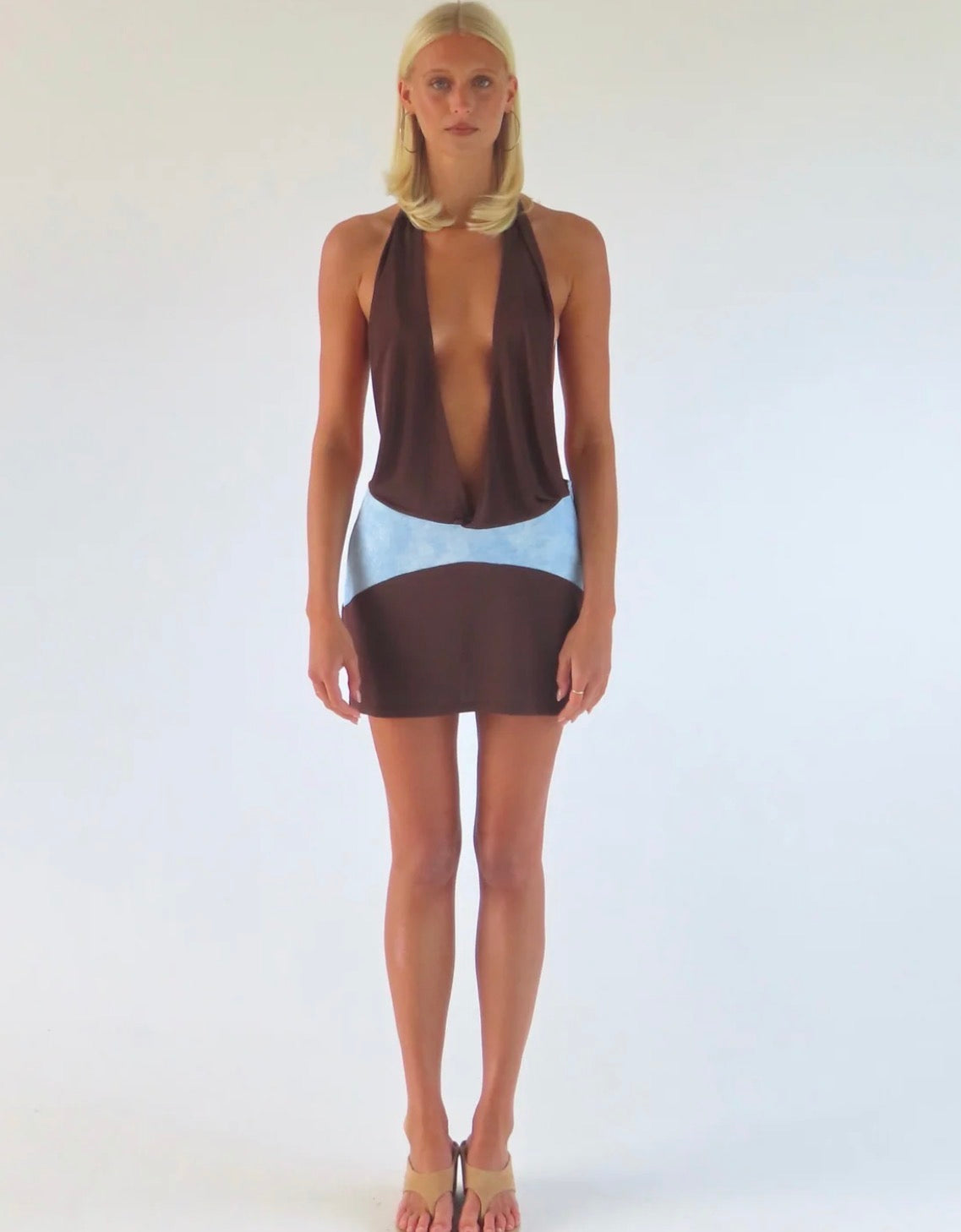  Model wearing the Sola Musa Oma Mini Dress in Bruna, featuring a deep V brown halter neckline, open silhouette and contrasting sky blue curved drop-waist panel.
