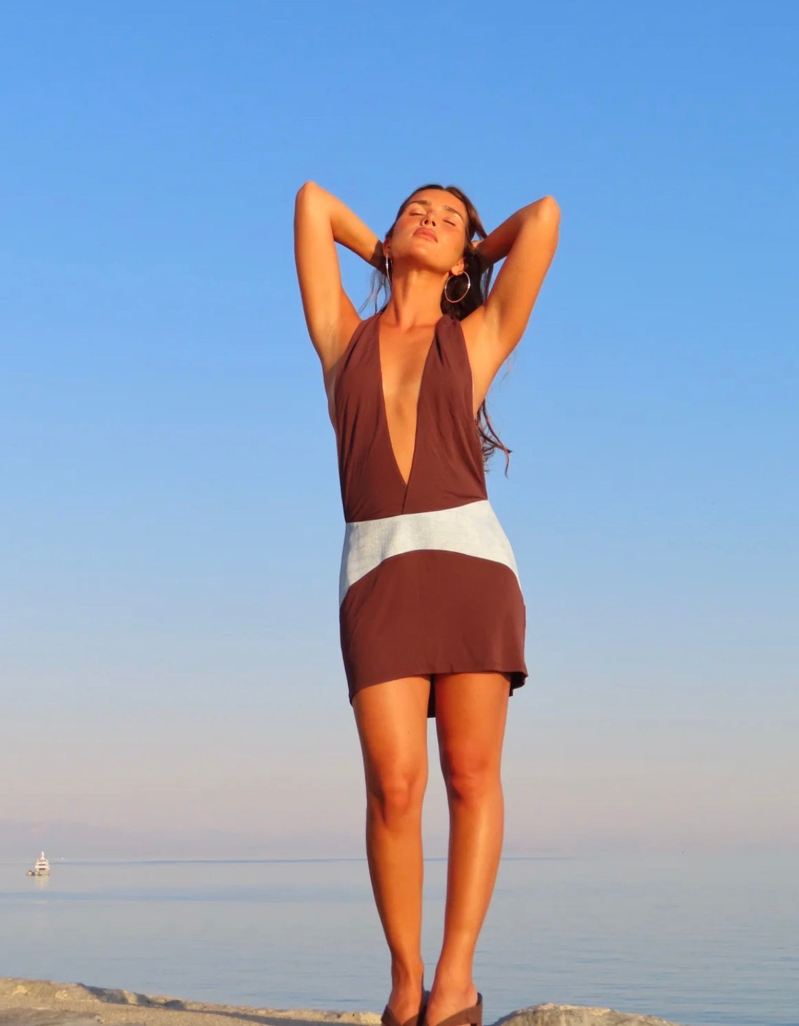  Model wearing the Sola Musa Oma Mini Dress in Bruna, featuring a deep V halter neckline, brown mini silhouette and contrasting sky blue curved drop-waist belt, photographed by the ocean.
