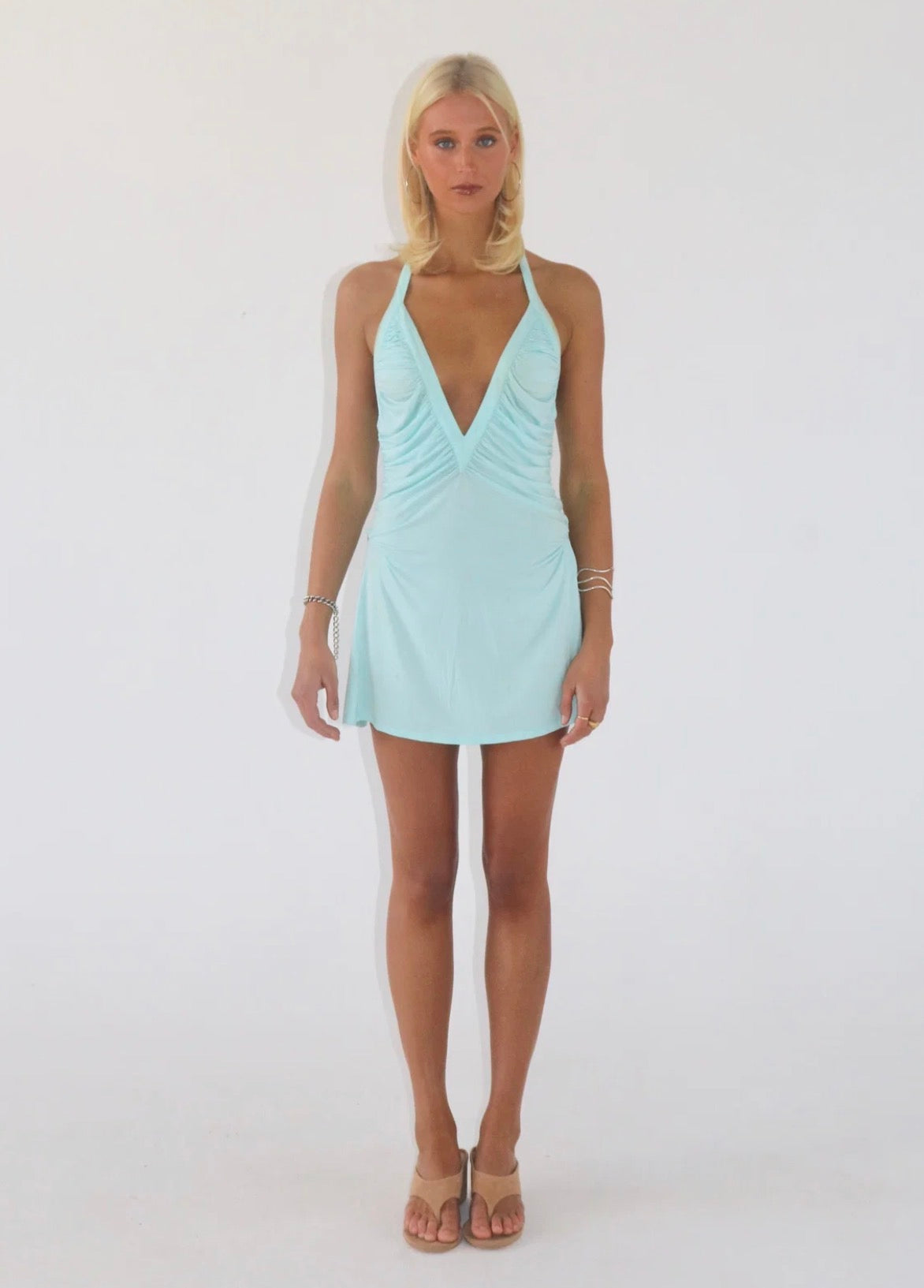 Model wearing the Angelina Mini Dress in light blue, featuring a deep V halter neckline, ruched bodice and flattering fitted mini silhouette.
