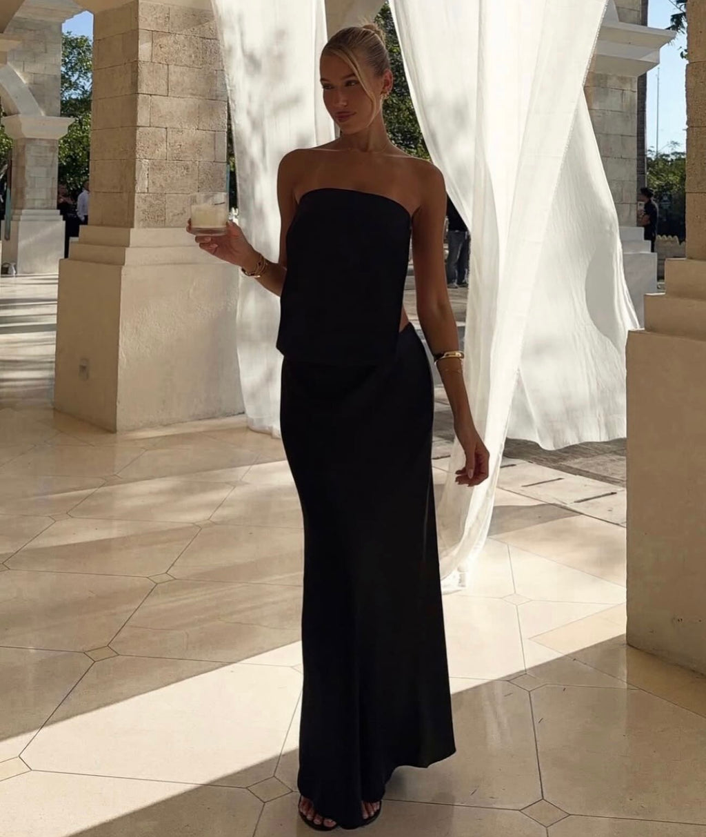 A woman models a strapless black maxi dress in an elegant outdoor colonnade with stone pillars and white drapes.