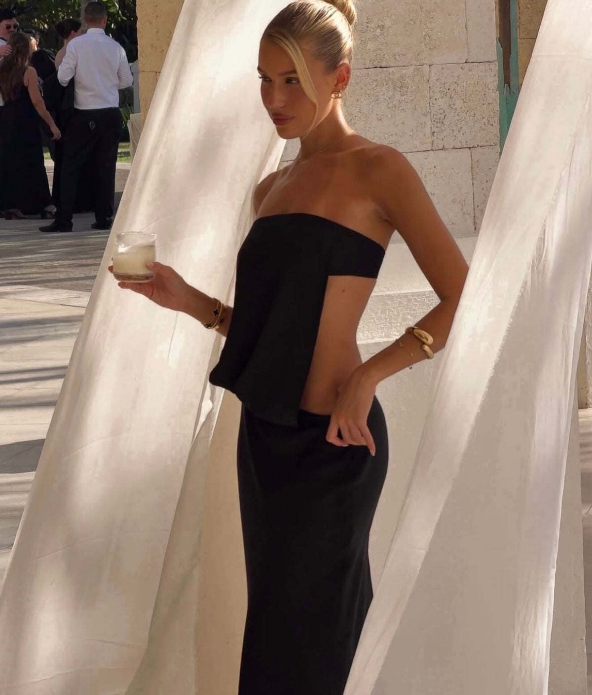 A woman wearing a black strapless peplum gown with side cutouts, holding a drink in a sunlit outdoor setting with sheer white drapes.