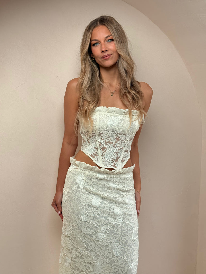 Woman wearing an ivory lace two-piece set featuring a strapless corset-style top with ruffle detailing and a matching lace midi skirt, styled with white pointed heels against a neutral studio background. - @indiabluegranger