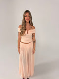A woman modeling a two-piece peach maxi set consisting of an off-the-shoulder ruched crop top and a matching pleated maxi skirt, set against a plain light gray studio background. - @sarahmadden