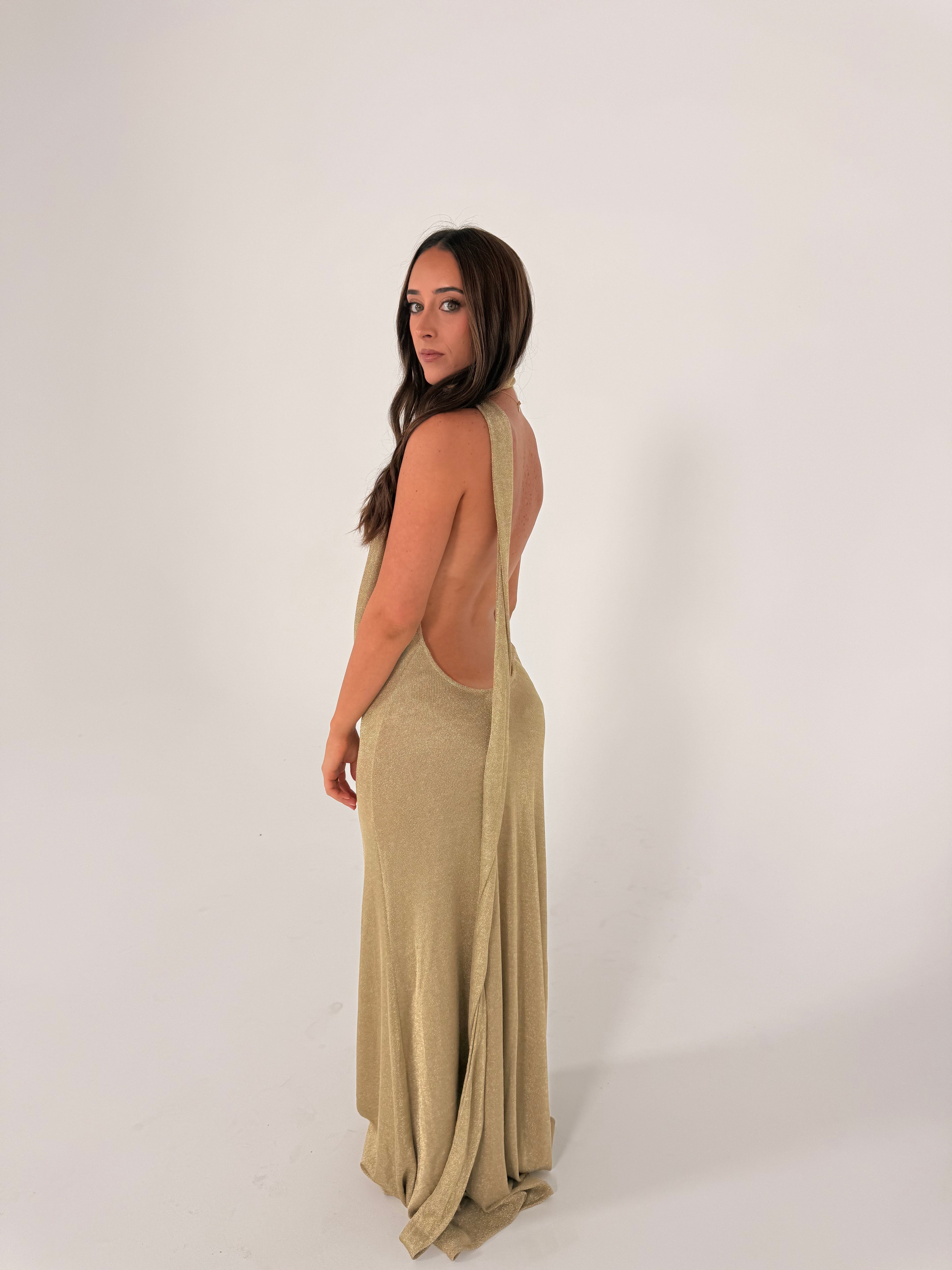 A woman modeling a sleeveless, high-neck, floor-length dress in a muted khaki-beige color, standing against a plain light gray studio background.
