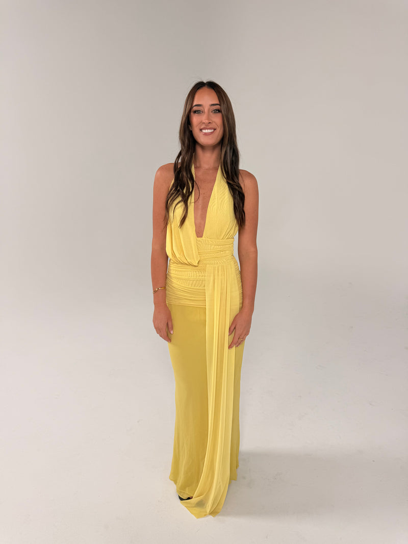 Front view of a woman wearing a pale yellow halter-neck maxi dress with a plunging neckline, ruched waist and long draped sash detail, photographed against a light studio backdrop. - @sarahmadden
