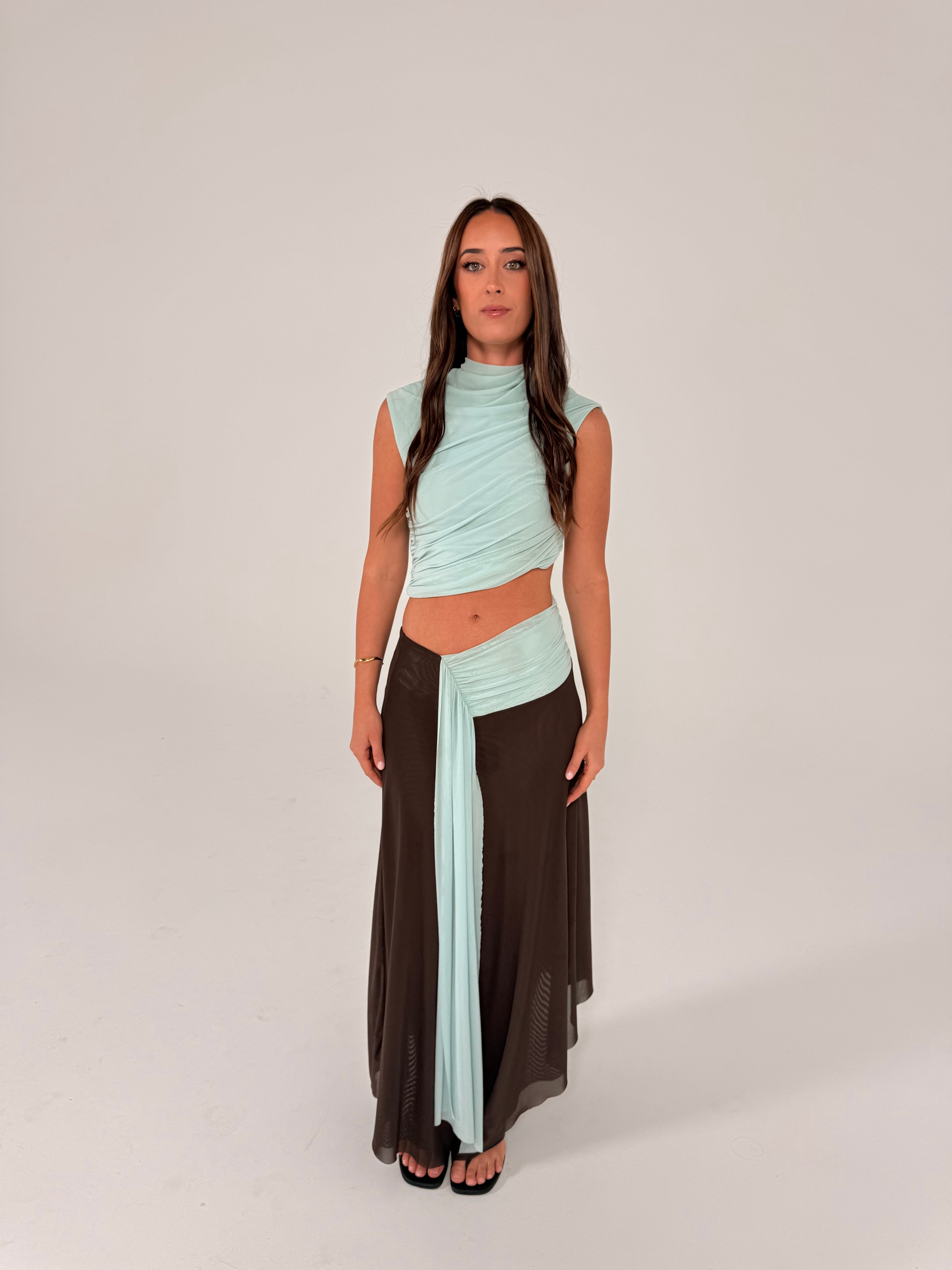 A woman modeling a two-piece outfit consisting of a light mint green wrap-style crop top and a long dark brown skirt, standing against a plain light gray studio background. - @sarahmadden
