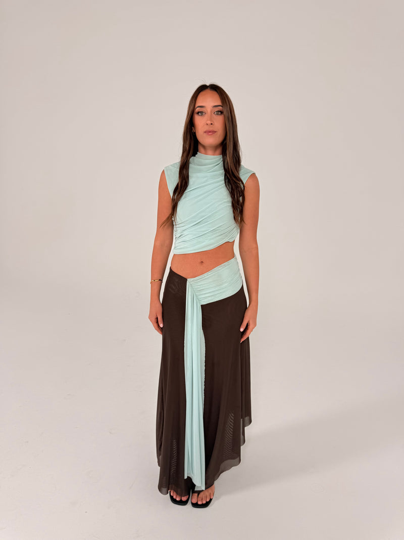 A woman modeling a two-piece outfit consisting of a light mint green wrap-style crop top and a long dark brown skirt, standing against a plain light gray studio background. - @sarahmadden