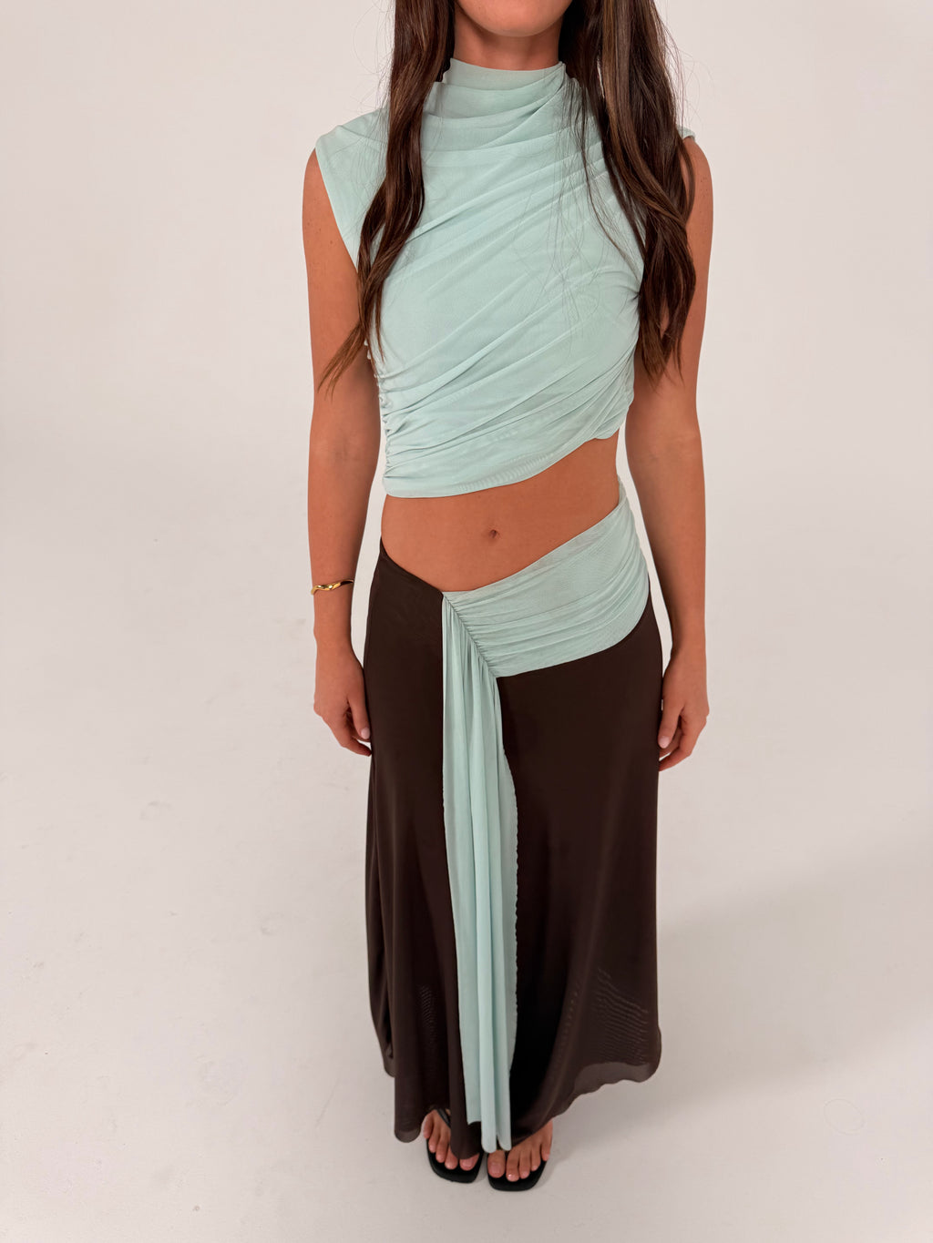 A woman modeling a two-piece outfit consisting of a light mint green wrap-style crop top and a long dark brown skirt, standing against a plain light gray studio background.