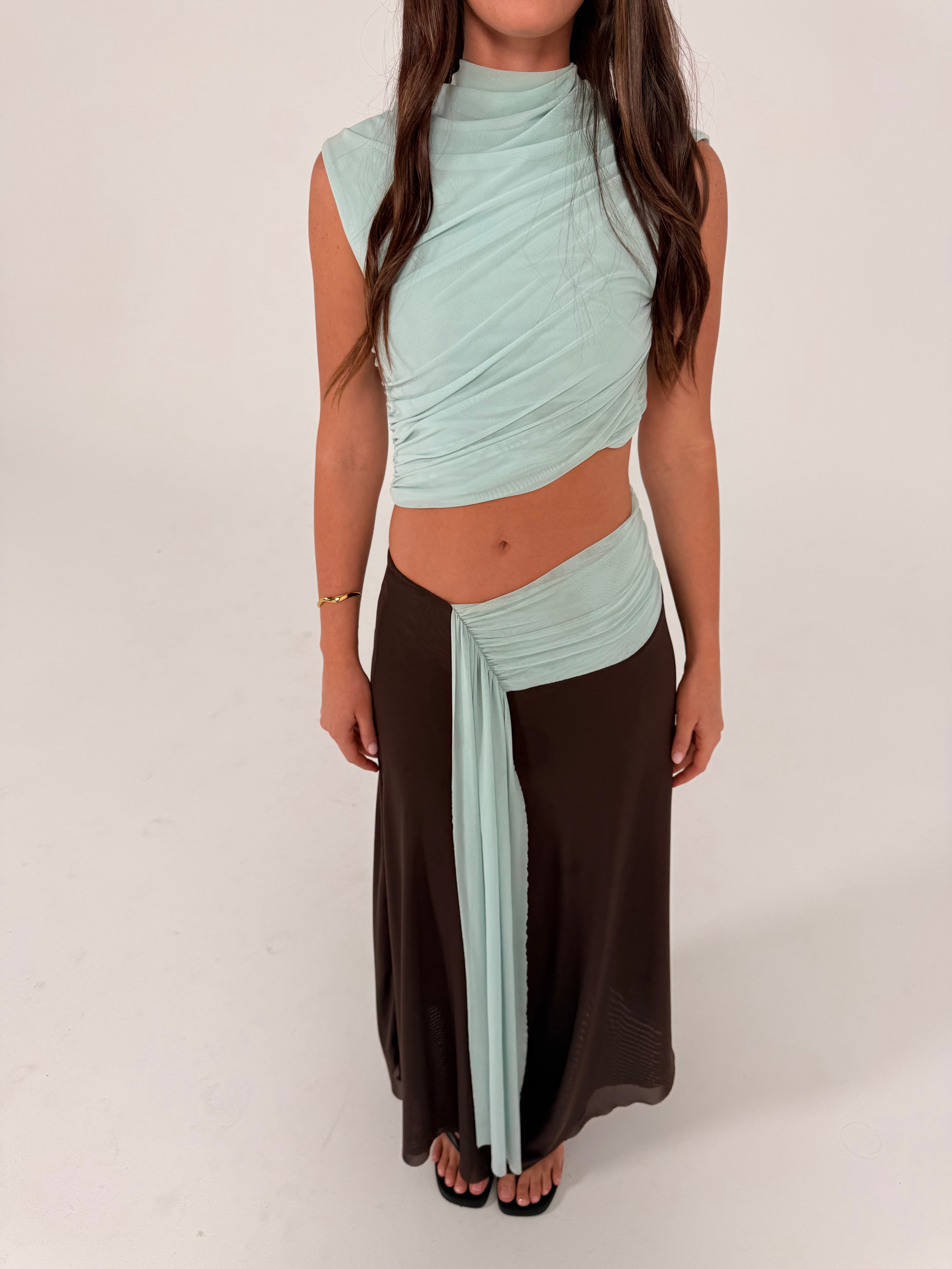 A woman modeling a two-piece outfit consisting of a light mint green wrap-style crop top and a long dark brown skirt, standing against a plain light gray studio background.