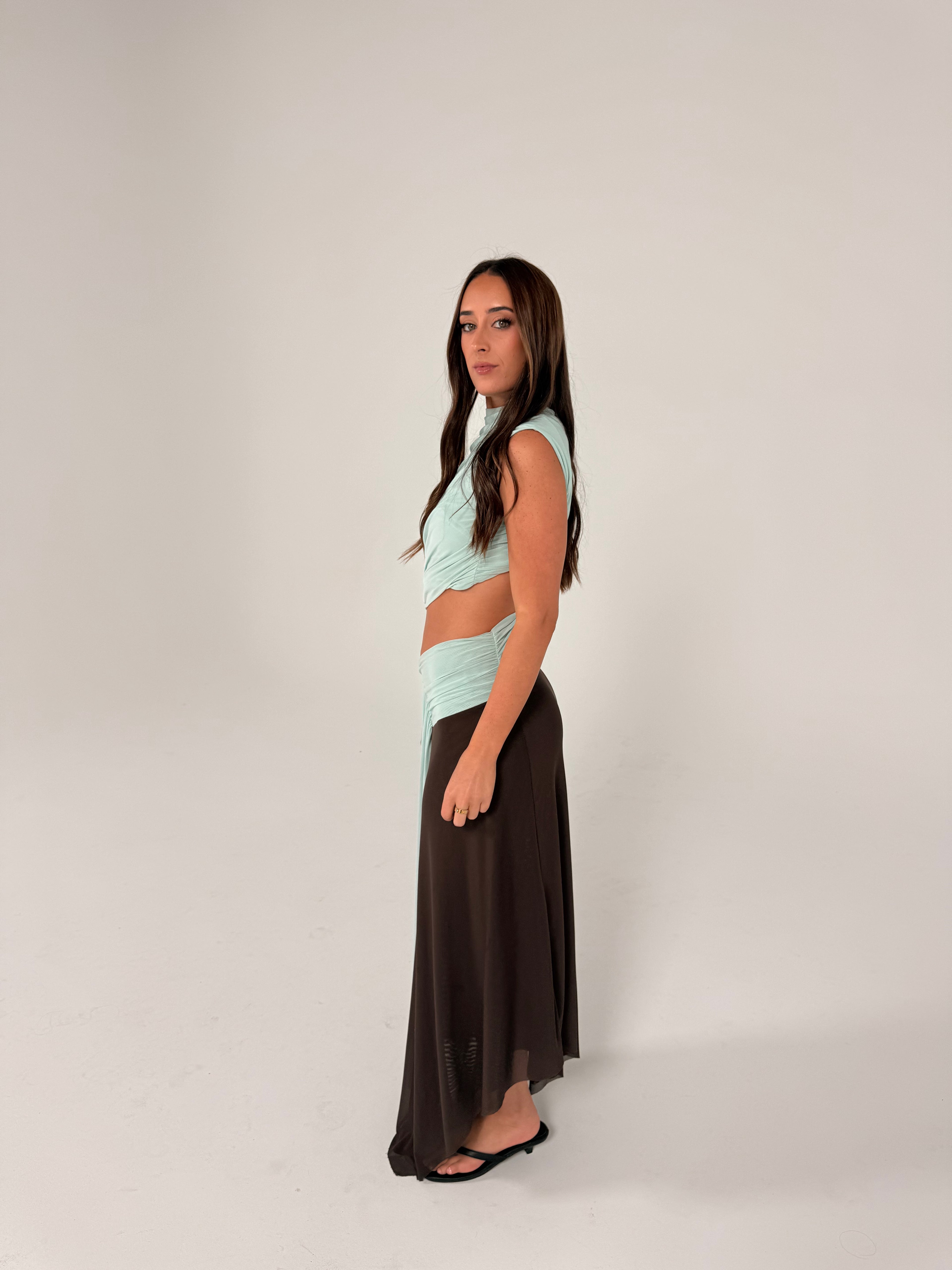 A woman modeling a two-piece outfit consisting of a light mint green wrap-style crop top and a long dark brown skirt, standing against a plain light gray studio background.