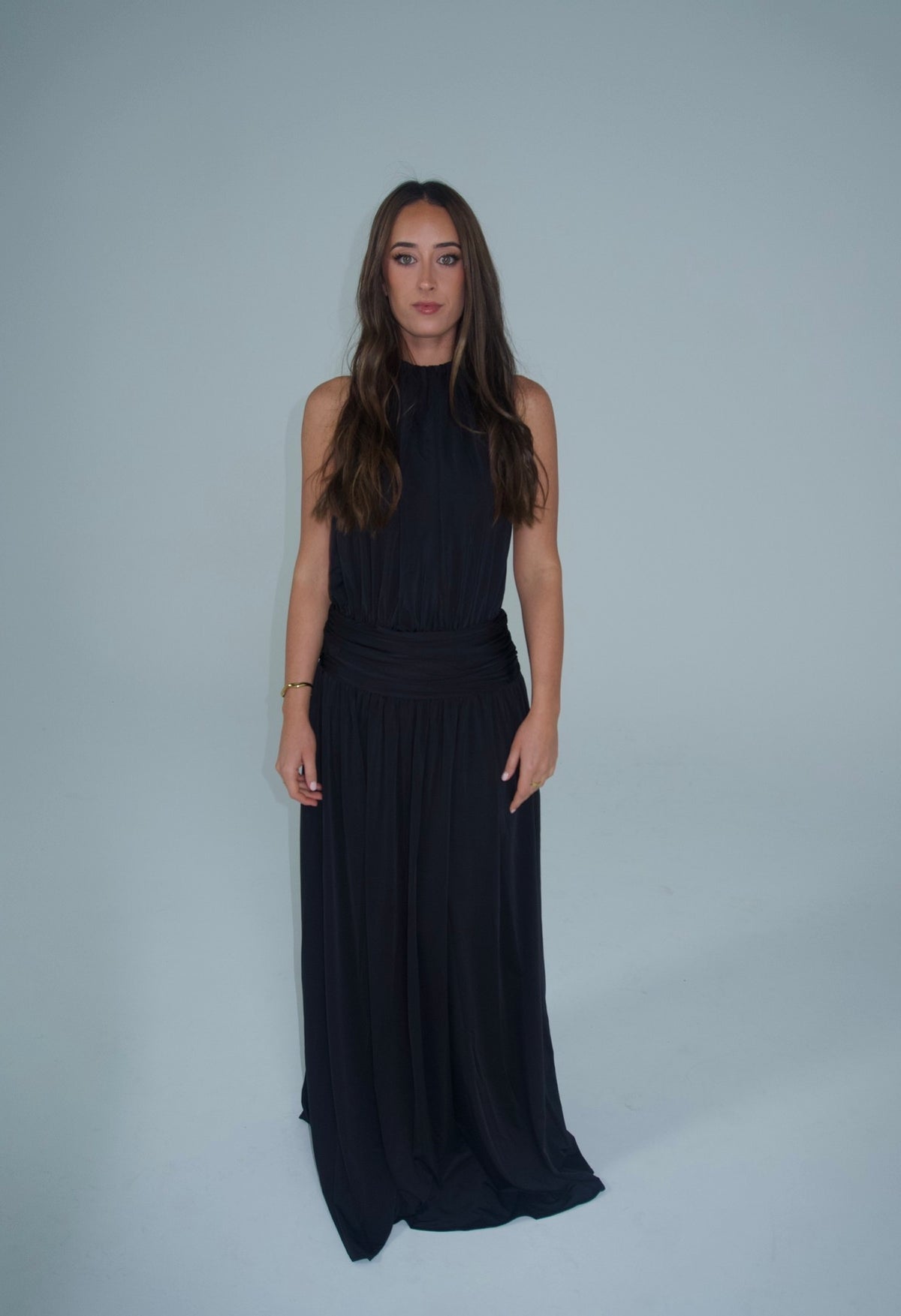 Front view of a woman wearing a black floor-length halter-neck maxi dress with a high neckline, softly gathered bodice, ruched waistband and flowing skirt, photographed against a light studio backdrop.