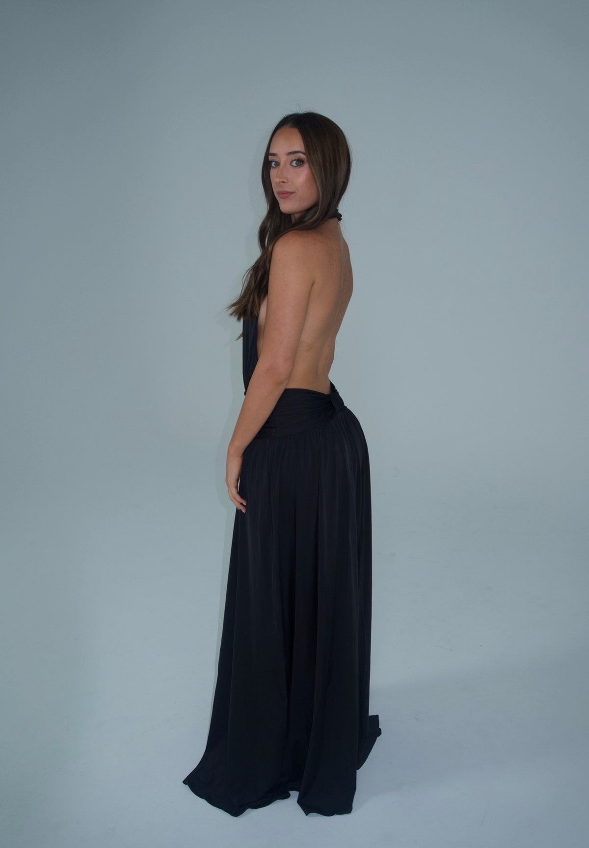 Side-back view of a woman wearing a black halter-neck maxi dress with a dramatic open back, low-waisted gathered detailing and a flowing floor-length skirt, photographed against a light studio backdrop.