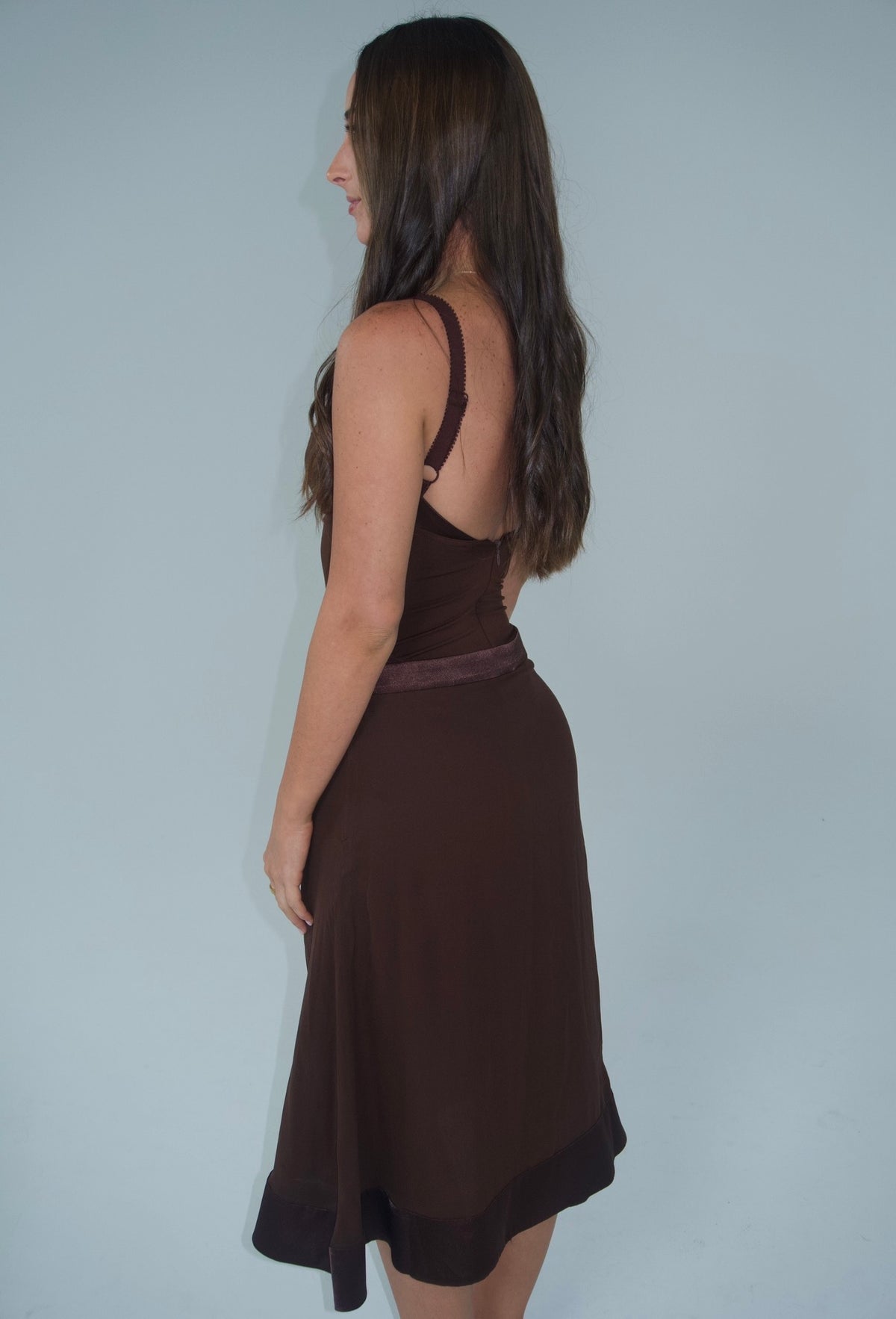 Side and back view of a woman wearing a chocolate brown fitted bustier top with adjustable straps and a matching flowing midi skirt with satin waistband and hem detailing, photographed against a light grey studio background.
