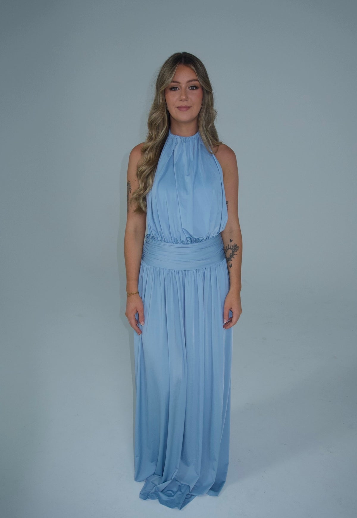 A woman modeling a light blue sleeveless halter-neck maxi dress with a matching fabric sash at the waist, standing against a solid light gray studio background. - @lilysulfaro