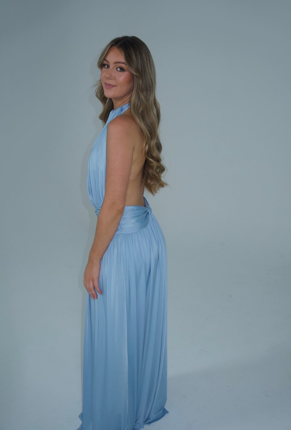 A woman modeling a light blue halter-neck maxi dress with a draped back and pleated skirt, standing against a plain light gray studio background.