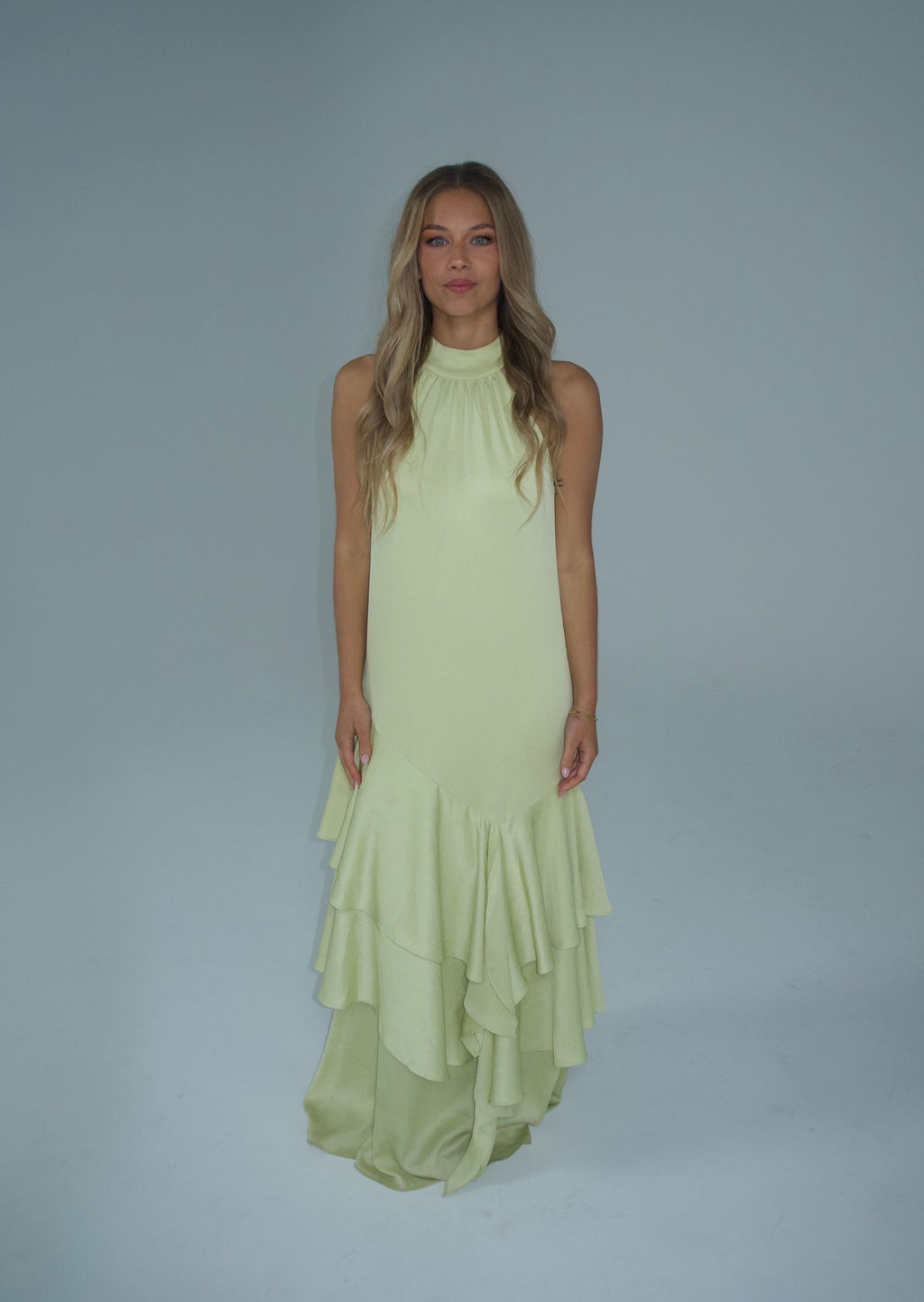 A woman modeling a sleeveless pale yellow-green halter-neck maxi dress with a tiered ruffled hem, set against a solid light gray studio background.