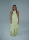 A woman modeling a sleeveless pale yellow-green halter-neck maxi dress with a tiered ruffled hem, set against a solid light gray studio background.