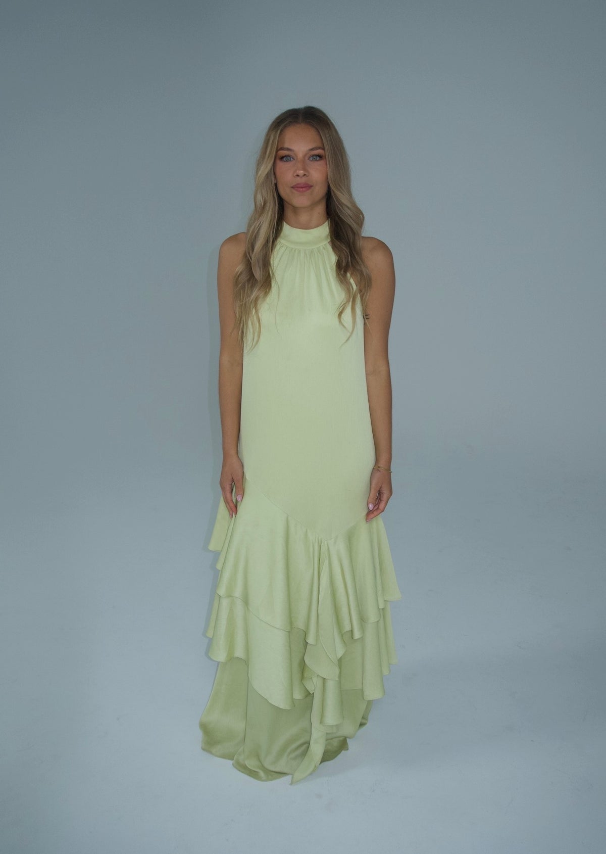 A woman modeling a sleeveless pale yellow-green halter-neck maxi dress with a tiered ruffled hem, set against a solid light gray studio background.