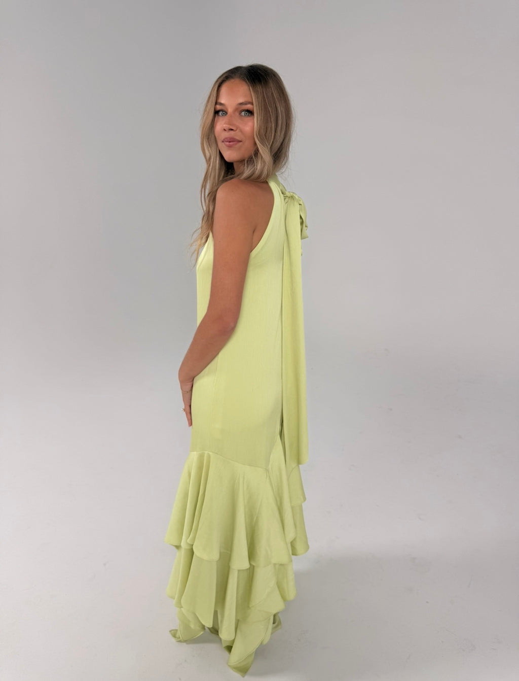 A woman modeling a sleeveless lime green maxi dress with a draped shoulder detail and a tiered ruffled hem, photographed against a neutral light gray studio background.
