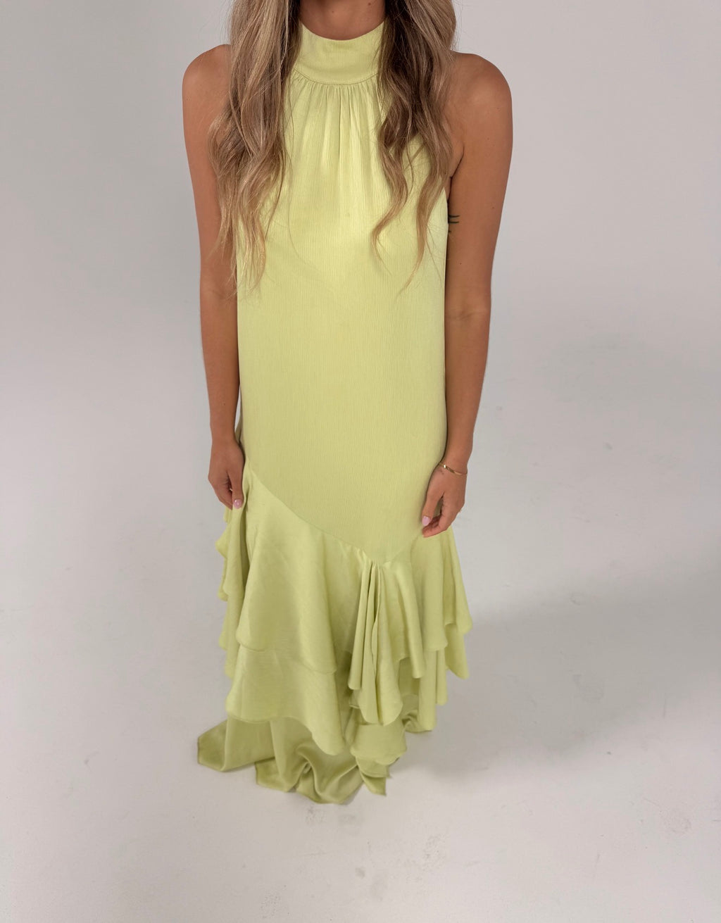 A woman modeling a sleeveless lime green maxi dress with a draped shoulder detail and a tiered ruffled hem, photographed against a neutral light gray studio background.