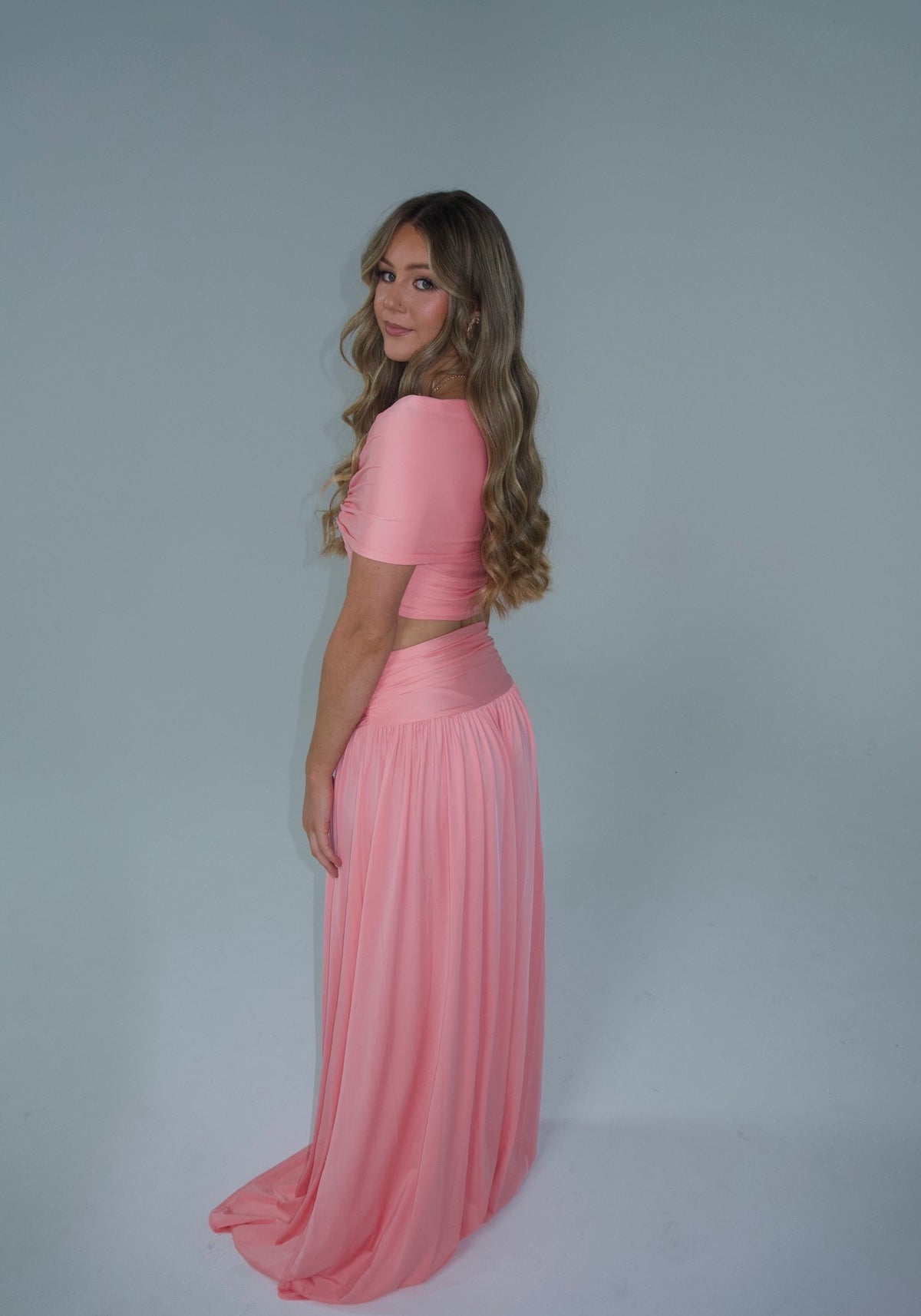 A woman modeling a light pink two-piece outfit consisting of a draped off-the-shoulder crop top and a matching long flowing skirt, set against a plain light gray studio background.