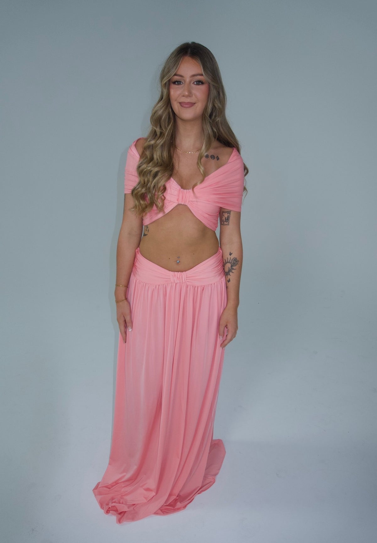 A woman modeling a light pink two-piece outfit consisting of a draped off-the-shoulder crop top and a matching long flowing skirt, set against a plain light gray studio background.