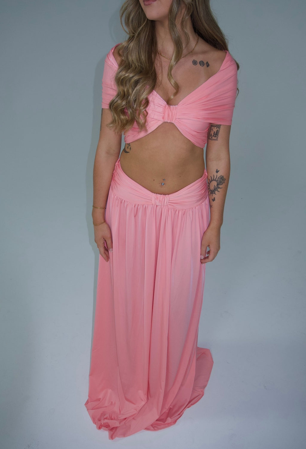 A woman modeling a light pink two-piece outfit consisting of a draped off-the-shoulder crop top and a matching long flowing skirt, set against a plain light gray studio background.