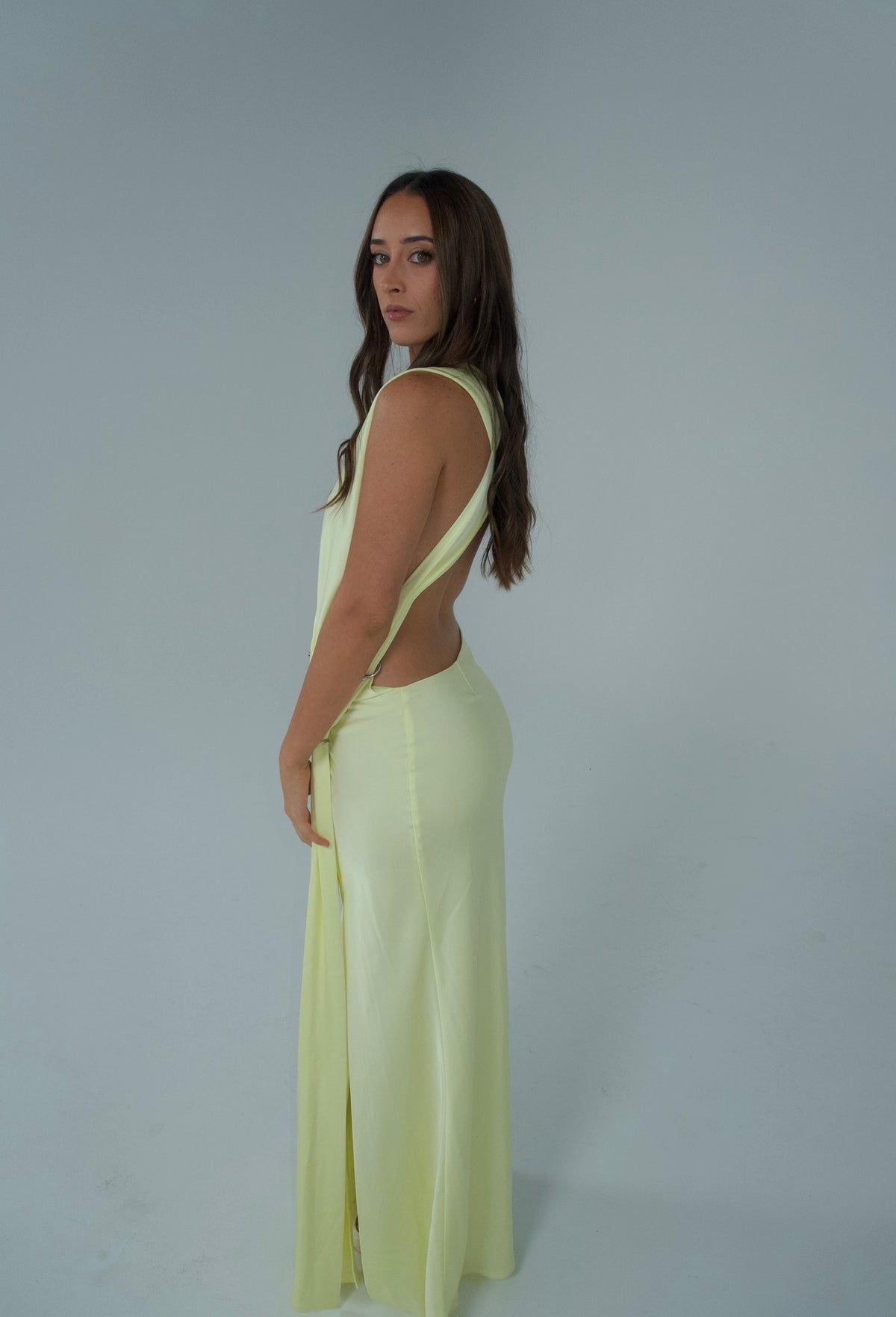 A woman modeling a sleeveless, pale yellow wrap-style maxi dress with a deep V-neckline, standing against a plain light gray studio background.