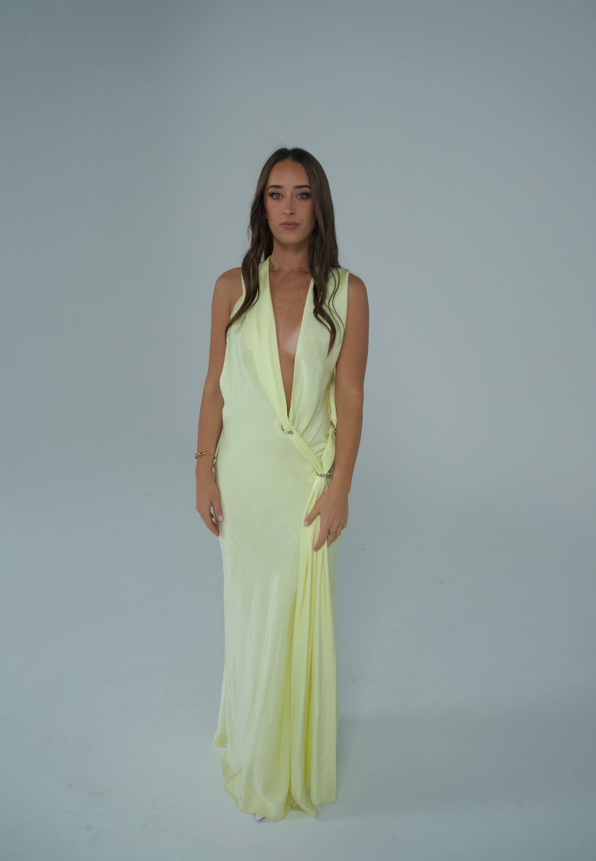 A woman modeling a sleeveless, pale yellow wrap-style maxi dress with a deep V-neckline, standing against a plain light gray studio background. - @sarahmadden