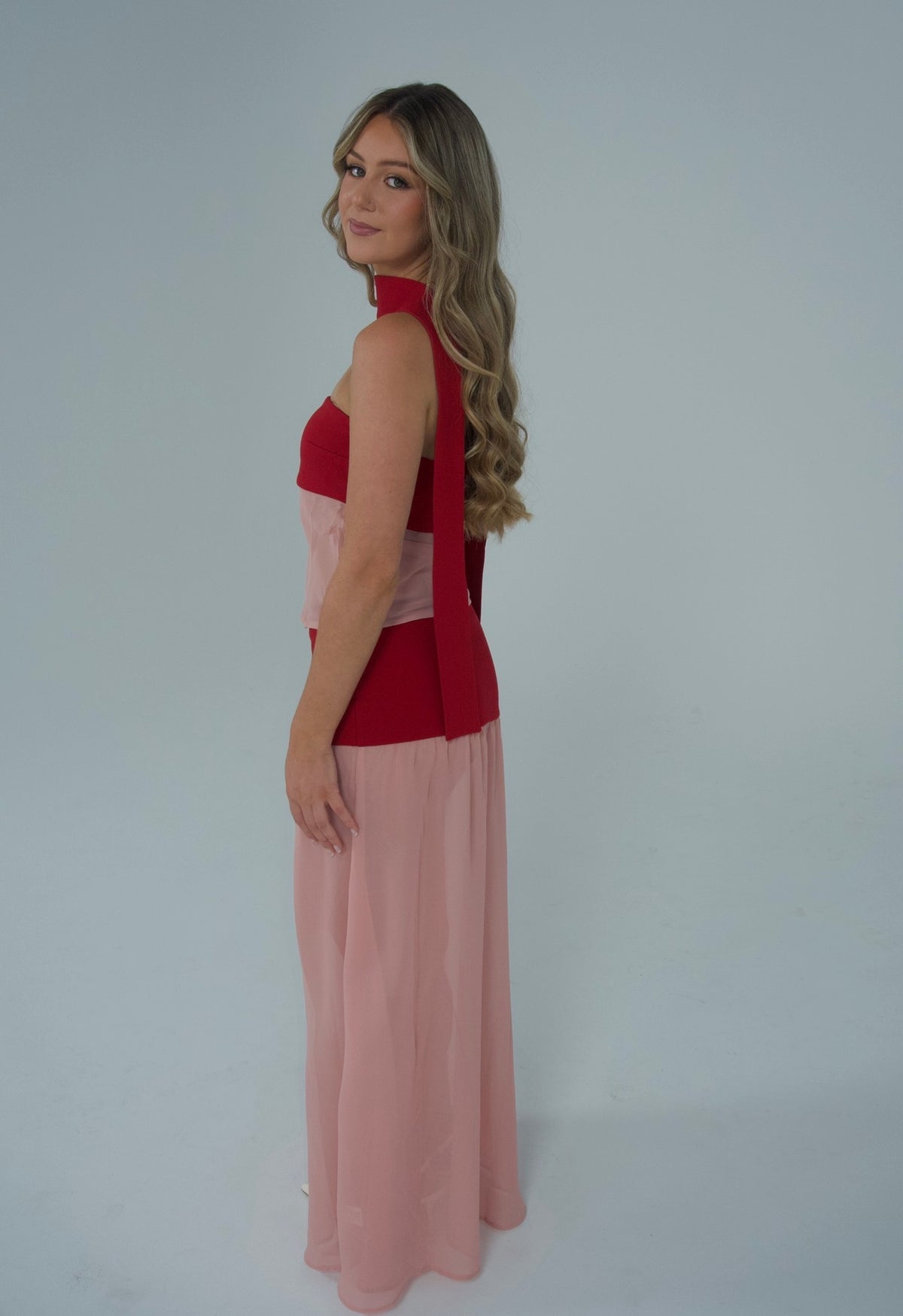 A woman modeling a sleeveless maxi dress with a red halter-style bodice and a flowing light pink skirt, photographed against a plain light gray studio background.