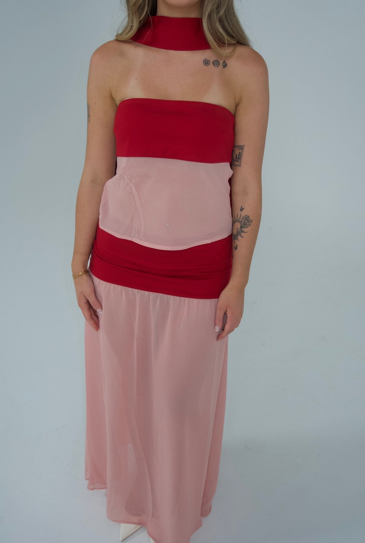 A woman modeling a sleeveless maxi dress with a red halter-style bodice and a flowing light pink skirt, photographed against a plain light gray studio background.
