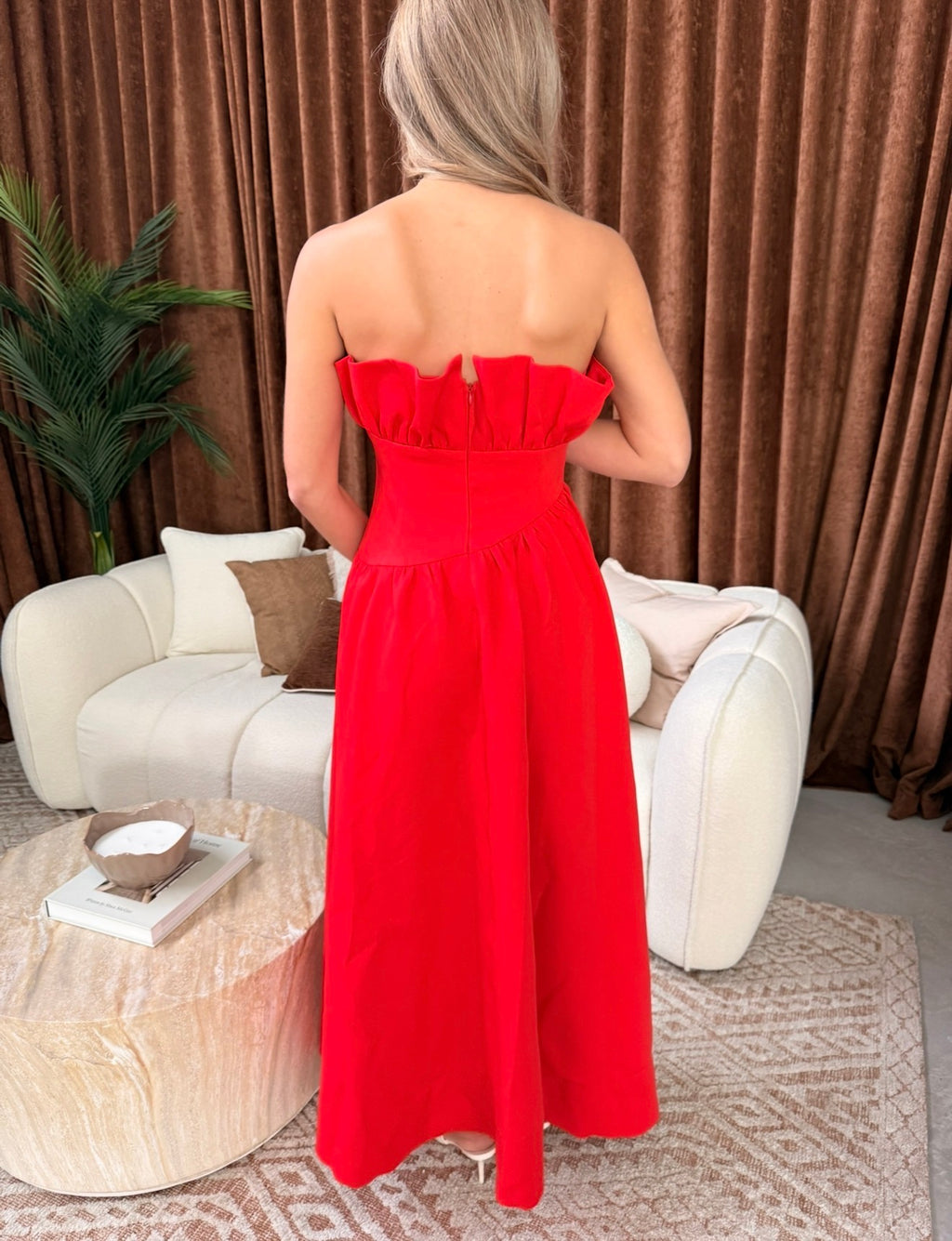 A woman stands with her back to the camera wearing a bright red strapless maxi dress with a ruffled neckline in a styled living room setting with a white sofa and brown curtains.