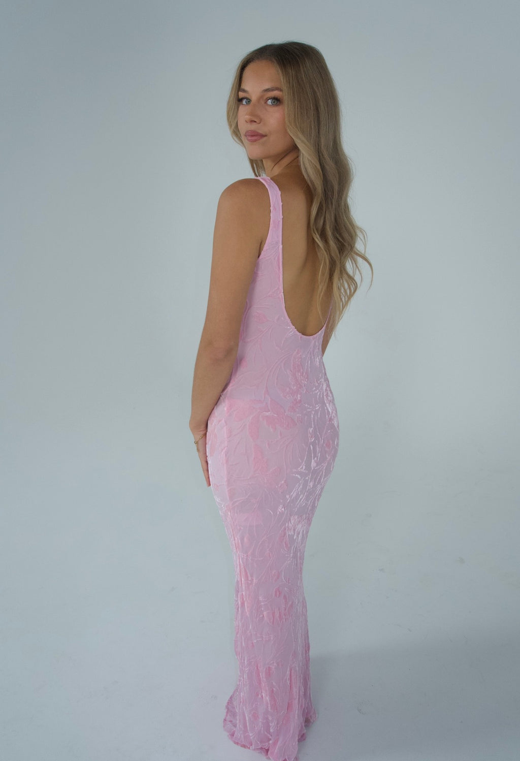 A woman modeling a light pink sleeveless lace maxi dress with a deep open back, photographed against a plain light gray studio background.