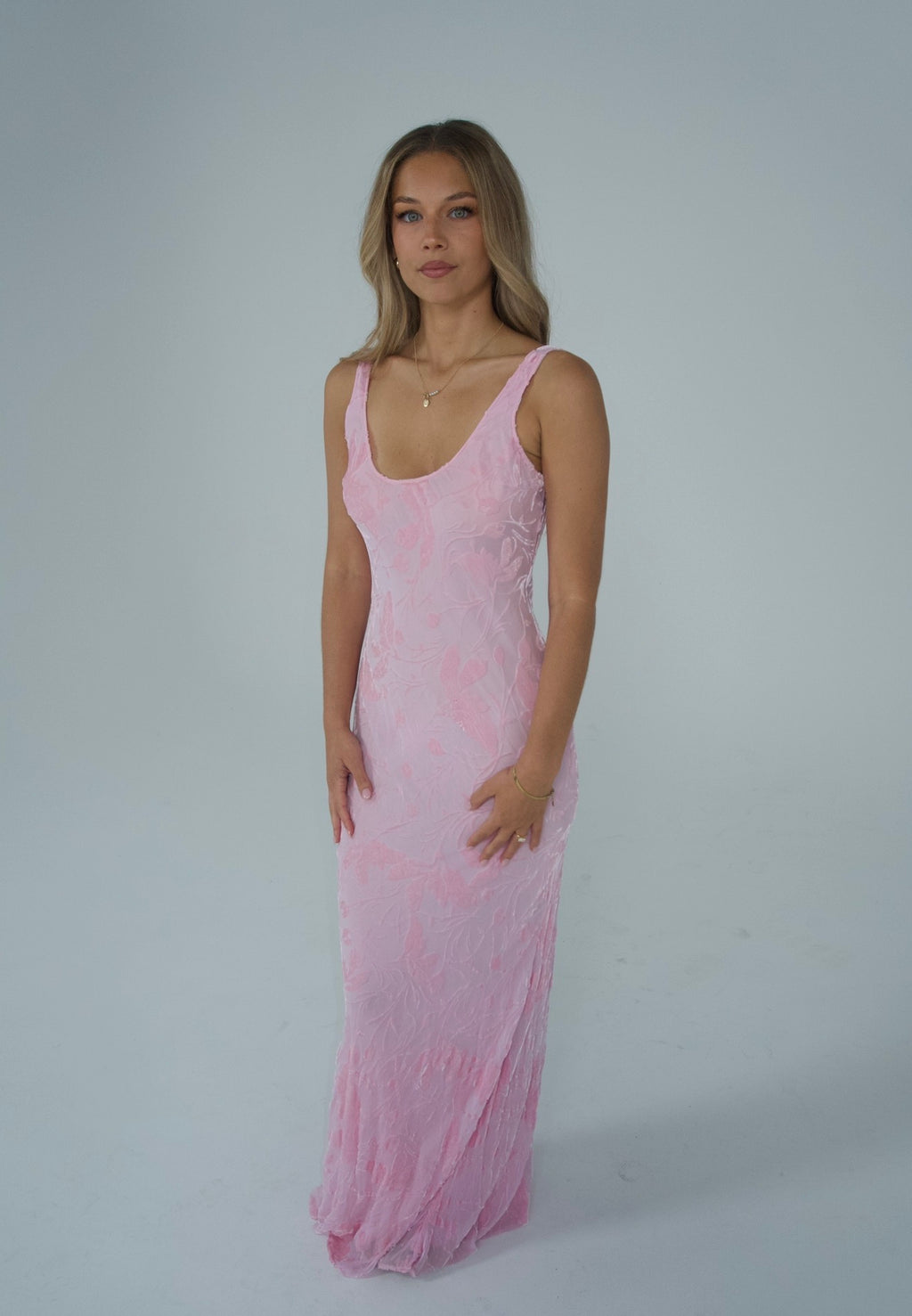 A woman modeling a light pink sleeveless maxi dress with a textured, sheer lace-like pattern against a plain light gray studio background.