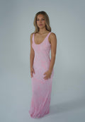 A woman modeling a light pink sleeveless maxi dress with a textured, sheer lace-like pattern against a plain light gray studio background.