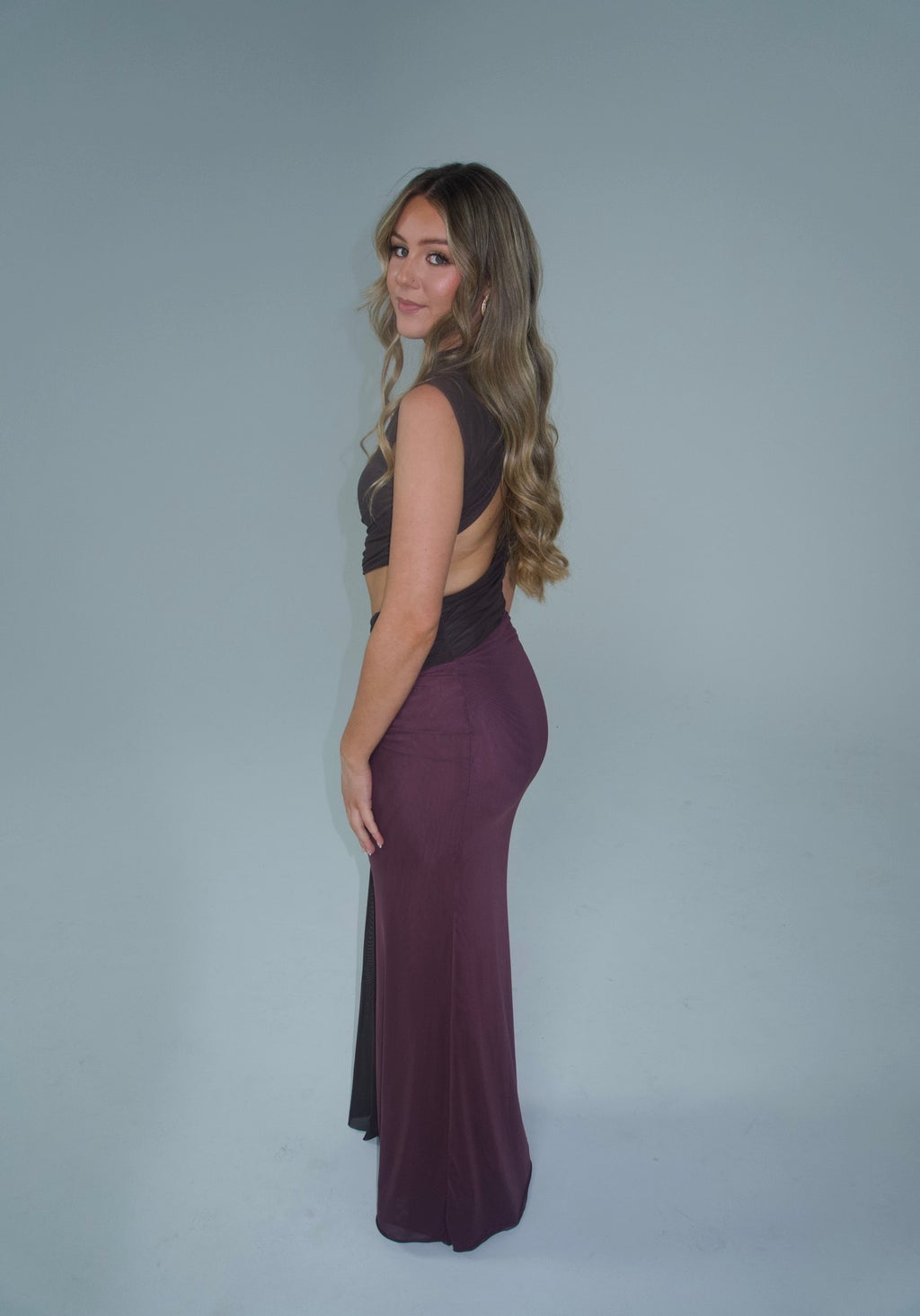A woman modeling a sleeveless, backless dark brown crop top and a matching deep plum purple maxi skirt, standing against a solid light gray studio background. - @lilysulfaro